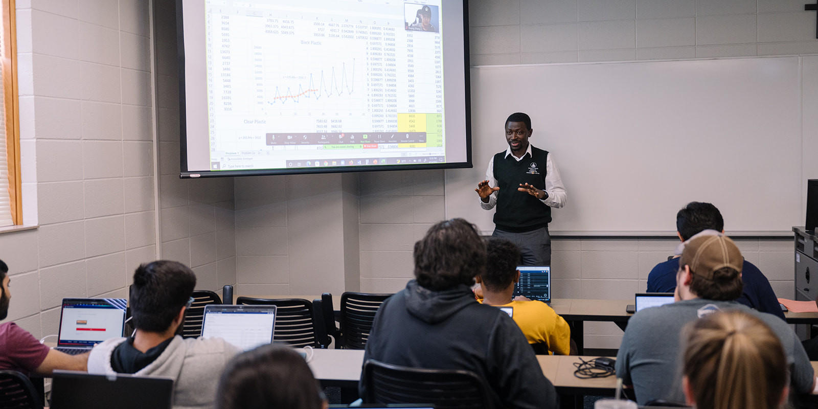 An instructor speaks with arms outstretched to seated students, next to a projection screen showing an Excel document being shared over Zoom.